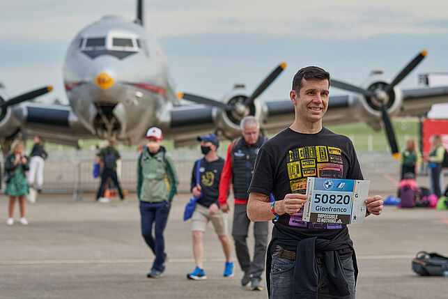 A young man holds his race number and beams into the camera. Behind him you can see an airplane.
