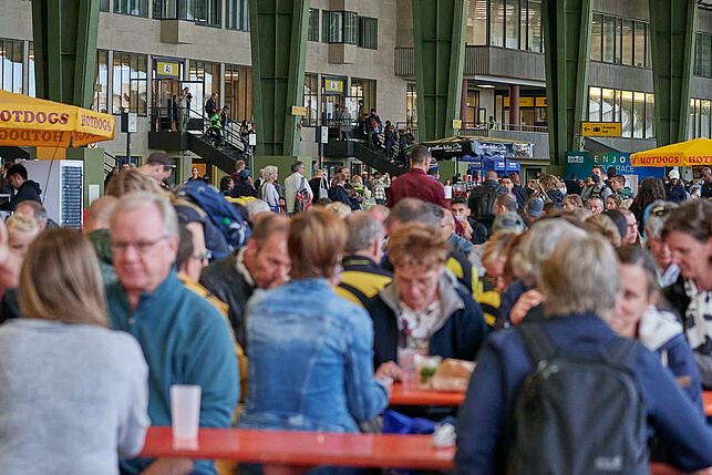 Auf dem sonnigen Flugfeld vor den Messehallen sitzen viele Menschen an Tischen, essen und unterhalten sich.
