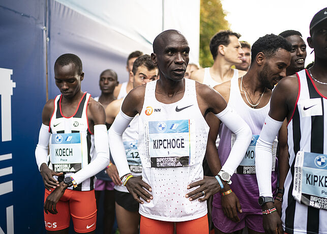 Weltrekordhalter Eliud Kipchoge steht konzentriert an der Startlinie und ist von zwei Tempomachern umgeben.