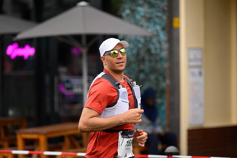 Man wearing sunglasses and a baseball cap runs at the event “Die Generalprobe”; he is wearing a vest with water bottles.