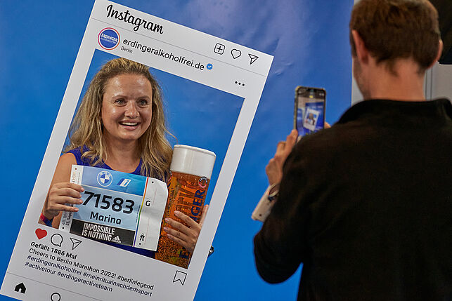 Eine glücklich lachende Frau macht ein Erinnerungsfoto für Instragram mit ihrer Startnummer im Fotorahmen von Erdinger.