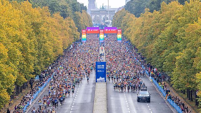 Shortly after the starting signal, thousands of runners take to the course and pass the starting gate in rainbow colors. At the very front you can see the top of the field behind the lead car.