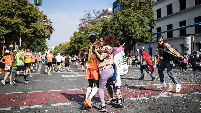 On the group, two runners are embraced by two women, beaming with joy. Behind them, many other runners are running.