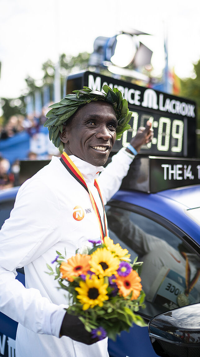 Eliud Kipchoge points to the record time displayed on the roof of the lead car. He is wearing a laurel wreath.