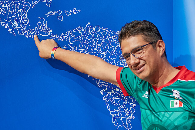 Vor einer großen Karte, auf der unzählige Trainingsstrecken eingezeichnet sind, zeigt ein männlicher Teilnehmer auf sein Heimatland in Südamerika.