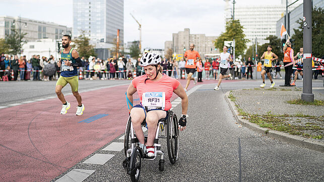 Some runners and a participant in a wheelchair come around the bend and turn into Karl-Marx-Allee.