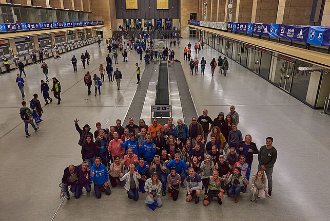 A group of people of different ages from all over the world are standing at the Marathon Fair in the main hall of Tempelhof Airport taking a group picture. Behind them you can see the baggage carousel.