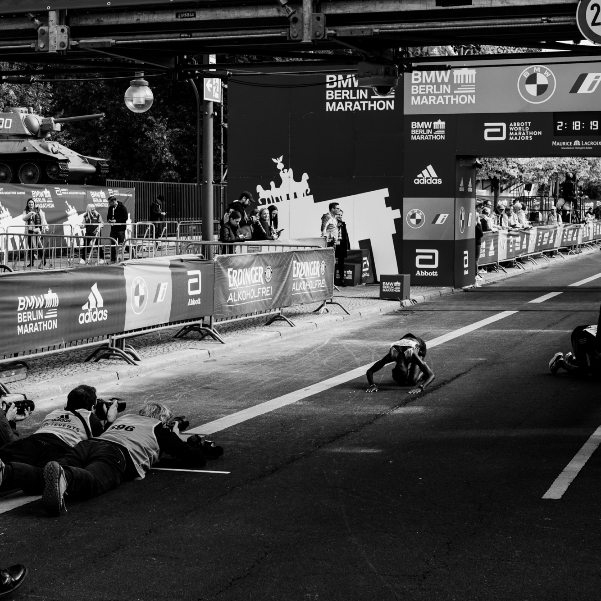 High-contrast black-and-white photo: elite runner behind finish line rests her forehead on the ground as if in prayer. Photographers are lying in front of her.