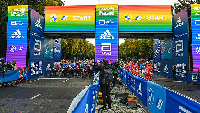 The start gate glows in rainbow colors. Photographers, participants and helpers run through the picture.