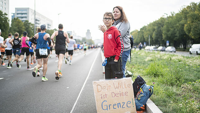 A woman and a child are standing at the track, in the foreground is a sign to cheer on the participants