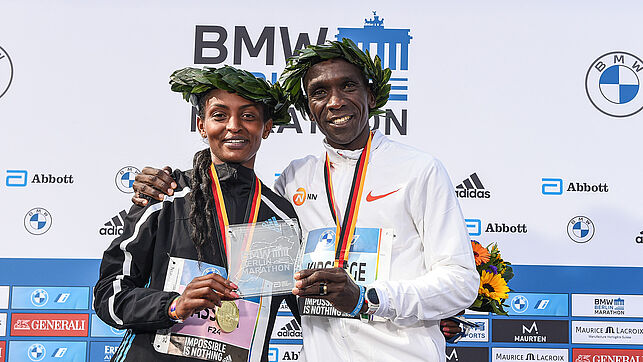 Eliud Kipchoge and Assefa Tigist are standing next to each other wearing laurel wreaths on their heads and holding a winner's trophy together. Kipchoge has his arm around Assefa.