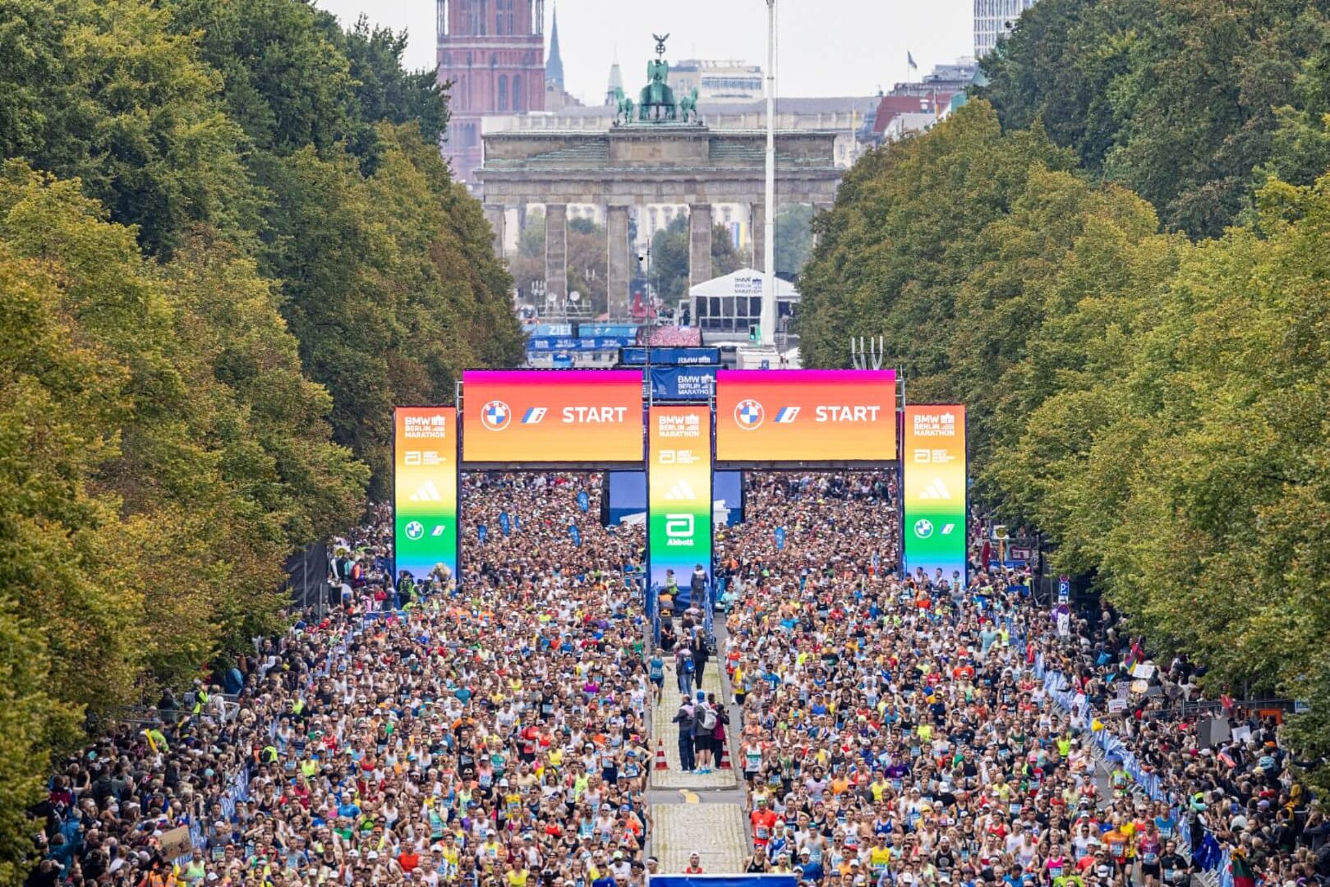 A huge crowd of runners passes through the starting gate, which is lit up in rainbow colours.