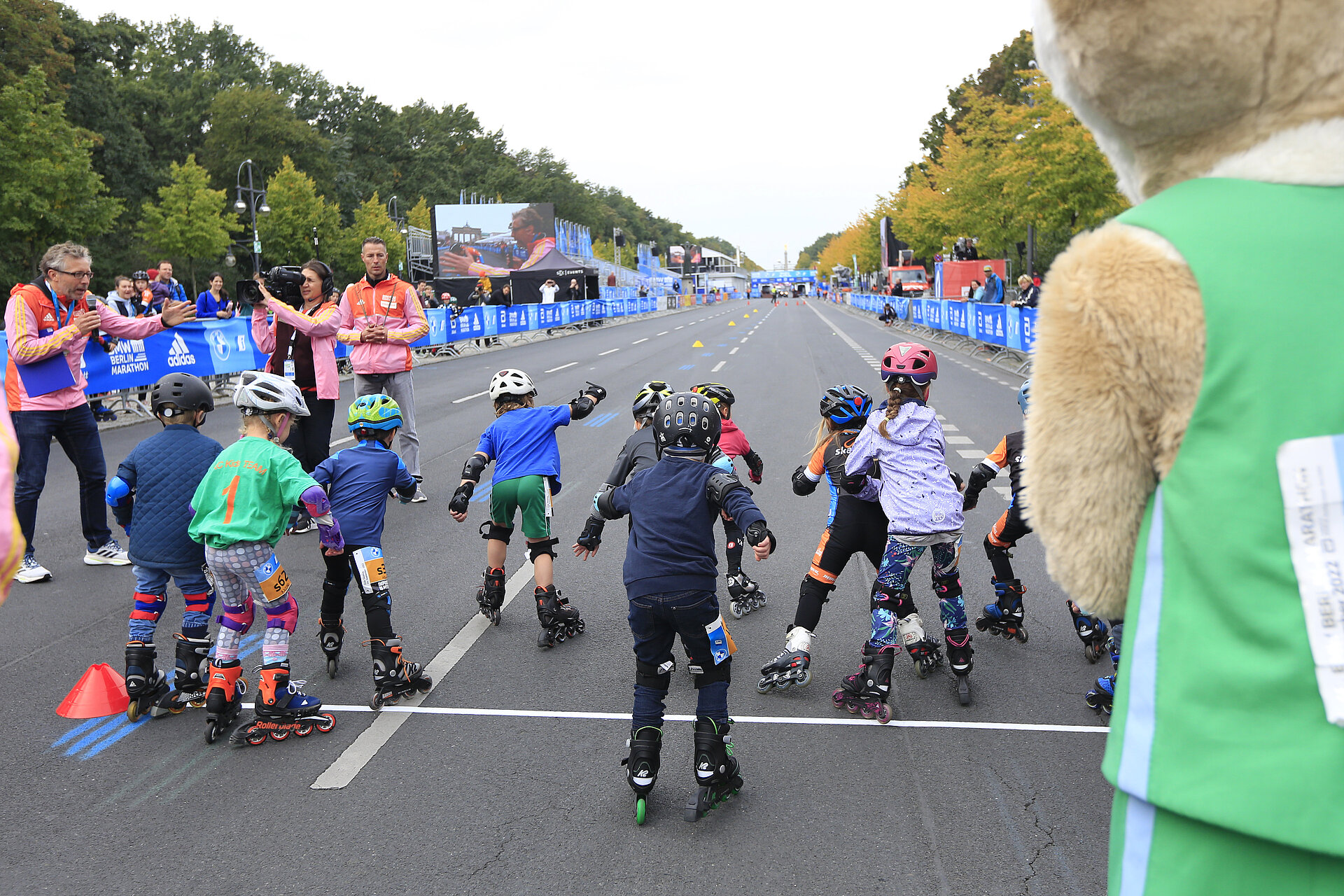 Kinder in Inline-Skates an der Startlinie auf einer gerade Straße. Zuschauende am Rand. SCC EVENTS Maskottchen Fridolin rechts im Vordergrund.