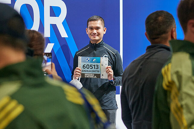 Ein mittelalter Mann posiert mit seiner Startnummer für ein Erinnerungsfoto auf der Marathon Expo und lächelt dabei glücklich.