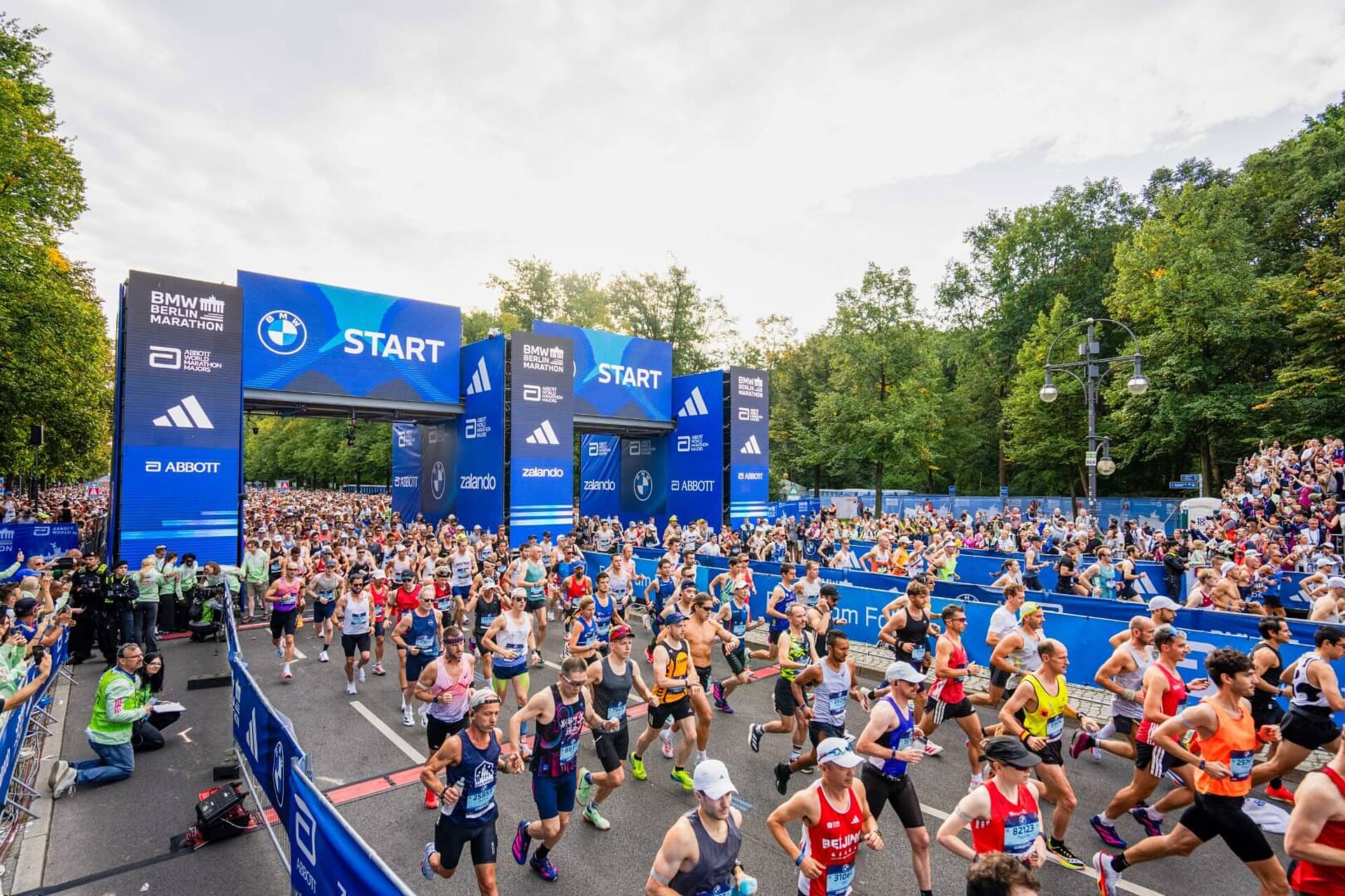 Runners pass through the starting gate and run past the course barriers, both of which feature sponsor logos.